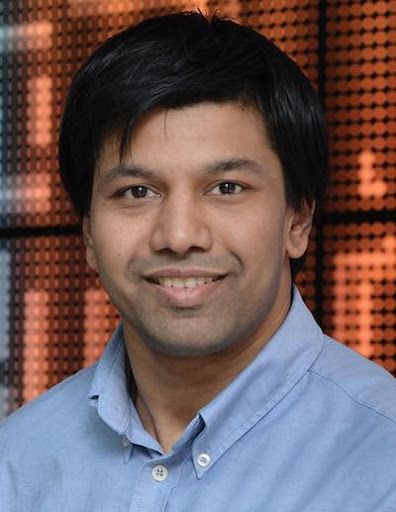 A headshot shows a smiling man with black hair wearing a light blue button-down shirt, with a blurred, orange and brown patterned background.
